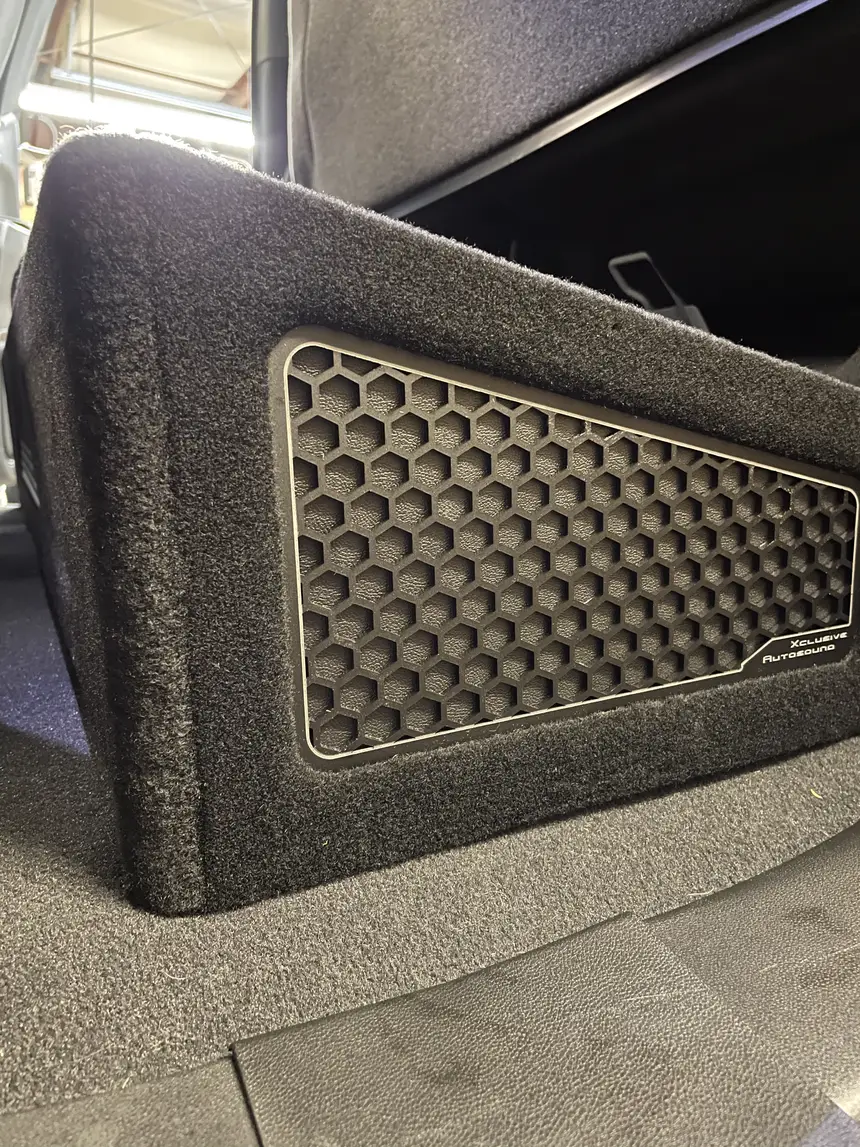Custom carpeted under-seat subwoofer enclosure with honeycomb grille and Xclusive Autosound badge in a truck, Belleville IL